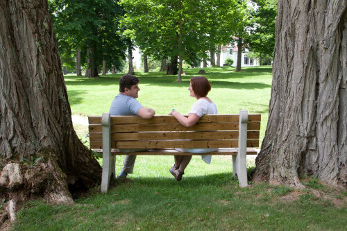 A man and a woman sit together on a park bench surrounded by huge trees, looking at one another and smiling